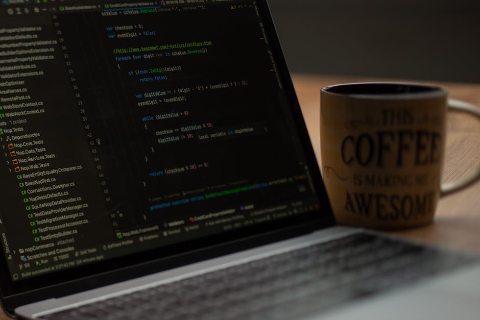 A close-up of a laptop displaying lines of code on its dark screen, placed on a wooden desk. Next to the laptop, there is a ceramic mug with a beige background and black text that says 'This coffee is