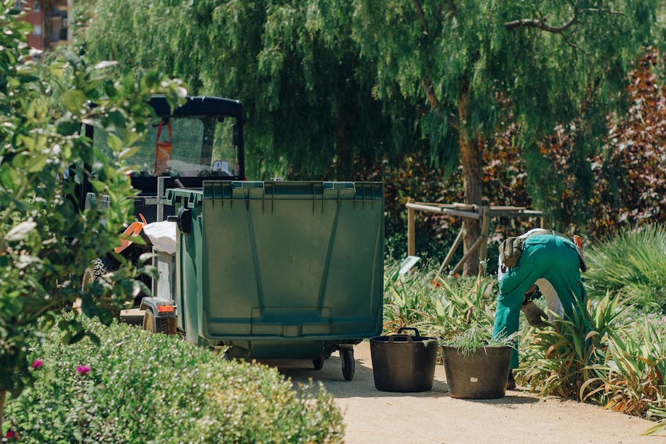 A gardener dressed in teal clothing and wearing a hat is crouched down on a garden pathway, actively trimming or planting in a bed of green, spiky-leaved plants. Nearby, two black containers sit on th
